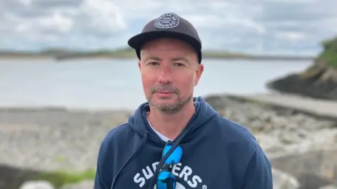 A man wearing a gray hoodie and a blue hoodie with sunglasses hung from the front of his hoodie standing by the sea