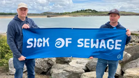 BBC Two men, both dressed in blue, stood by the sea holding a sign saying I'm sick from sewage